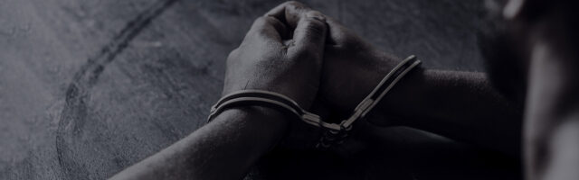 Close-up of a person's hands resting on a dark surface, with wrists secured in metal handcuffs, suggesting detention or arrest. The image has a somber, low-light atmosphere.