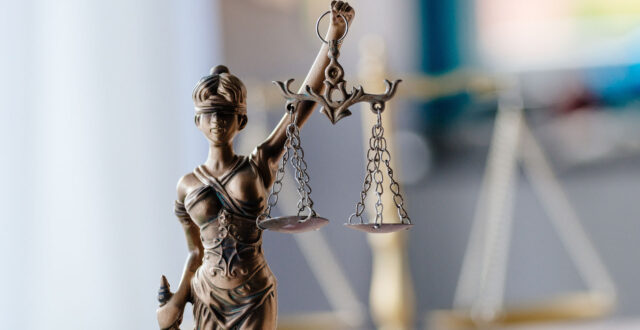 A close-up of a bronze Lady Justice statue holding a set of balance scales, with her eyes covered by a blindfold. The background is blurred, highlighting the statue as the main subject.