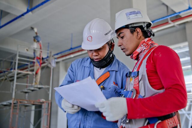 Two construction workers wearing safety helmets and gloves review a document together at an indoor construction site, ensuring everyone follows safety protocols so no one gets injured on the job. Scaffolding and exposed pipes are visible in the background.