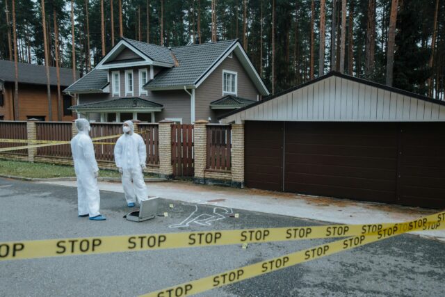 Two forensic investigators in protective suits examine a crime scene outside a house, marked with a chalk outline and evidence markers. Yellow 