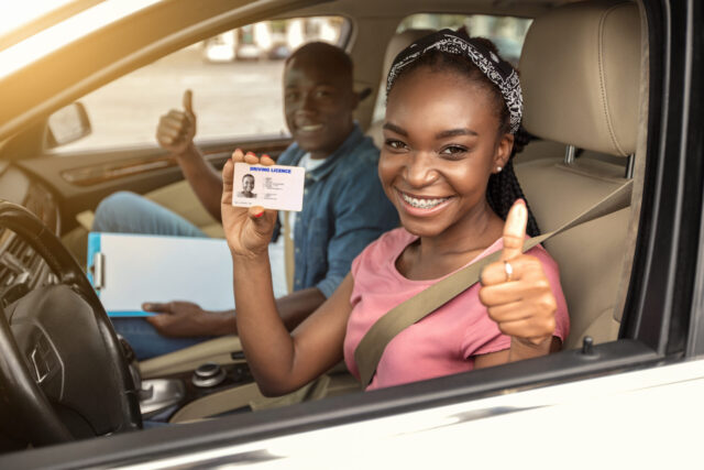 A smiling woman sitting in the driver’s seat of a car holds up her driver’s license and gives a thumbs-up. A man beside her, possibly an instructor, also gives a thumbs-up.