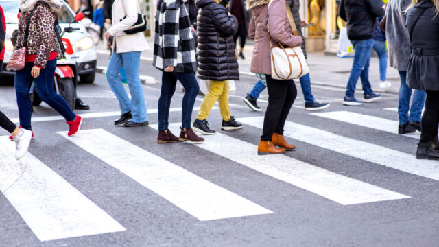 People wearing winter clothes walk across a crosswalk in a city. The scene is lively, with various coats, boots, and bags visible. The street and crosswalk lines are prominent in the foreground.