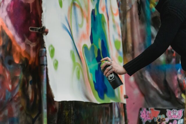 A person spray-paints colorful graffiti art with blue and green paint on a large canvas, surrounded by other vibrant painted surfaces. Only their arm and hand are visible.