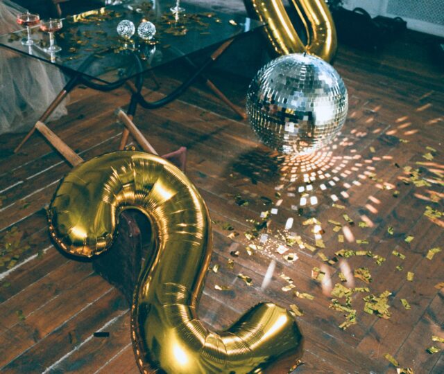 A shiny disco ball and large gold balloon shaped like the number 2 are on a wooden floor scattered with gold confetti, reflecting light. Glasses and decorations sit on a nearby glass table.