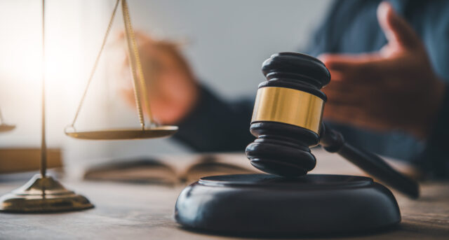 A wooden judge’s gavel rests on a sound block next to a balance scale on a desk, with a blurred person gesturing in the background, suggesting a legal or courtroom setting.