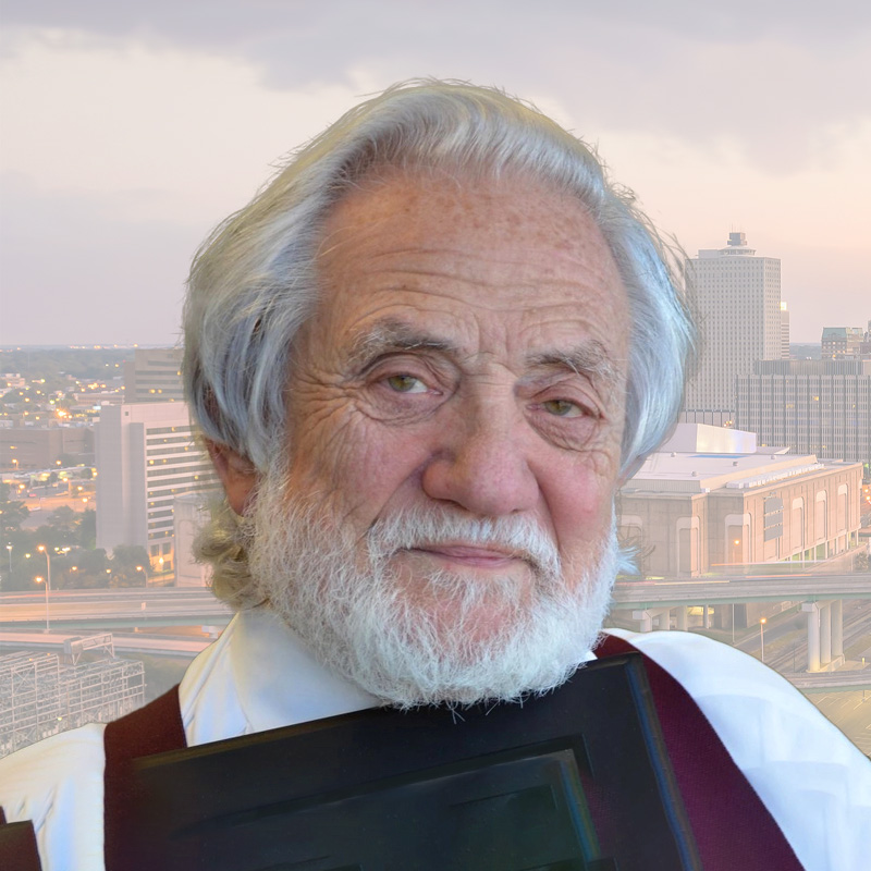 An elderly man with white hair and a beard smiles gently, standing in front of a cityscape with tall buildings and highways at sunset. He is holding a dark-colored object close to his chest.