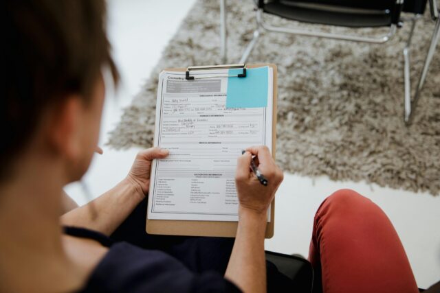A person holding a clipboard fills out a form with a pen. The scene appears to be in an office or counseling setting, with a carpet and empty chair visible in the background.