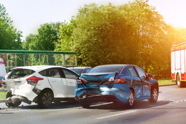 Two cars, a white one and a blue one, are involved in a rear-end collision on a sunlit road. Both vehicles have visible damage, and a fire truck is parked nearby. Trees and greenery are in the background.