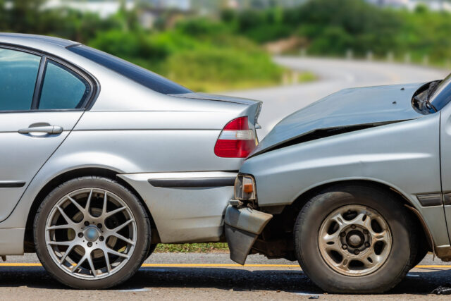A silver car has rear-ended another silver car on a road, damaging the front of one vehicle and the back of the other. The scene is outdoors with greenery and a curved road in the background.