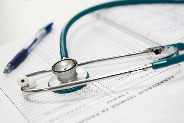 A stethoscope rests on top of a medical form with handwritten notes, next to a blue pen.