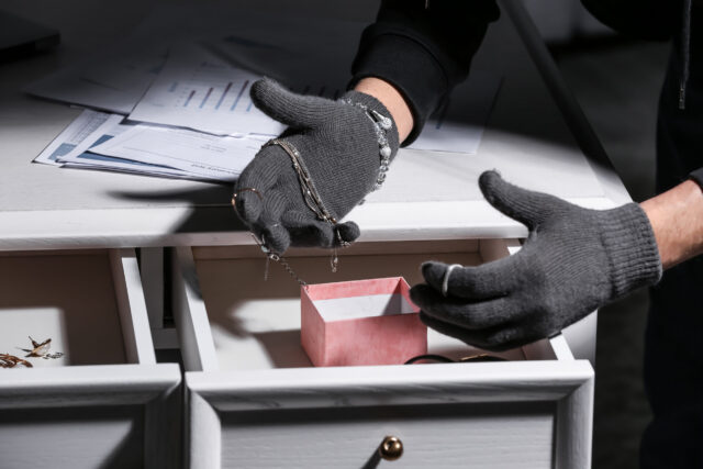 A person wearing gray gloves commits a theft felony by taking jewelry from an open drawer, with documents and a pink box also visible on the white table.