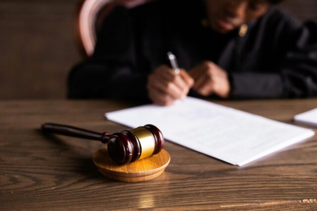 A judge wearing black robes writes on documents at a wooden desk, with a gavel and sound block in focus in the foreground, preparing paperwork related to a warrant search of property.